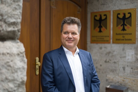 Ein Mann im Anzug steht lächelnd vor einer Holztür, daneben sind Schilder mit Bundesadlern an der Steinwand befestigt.