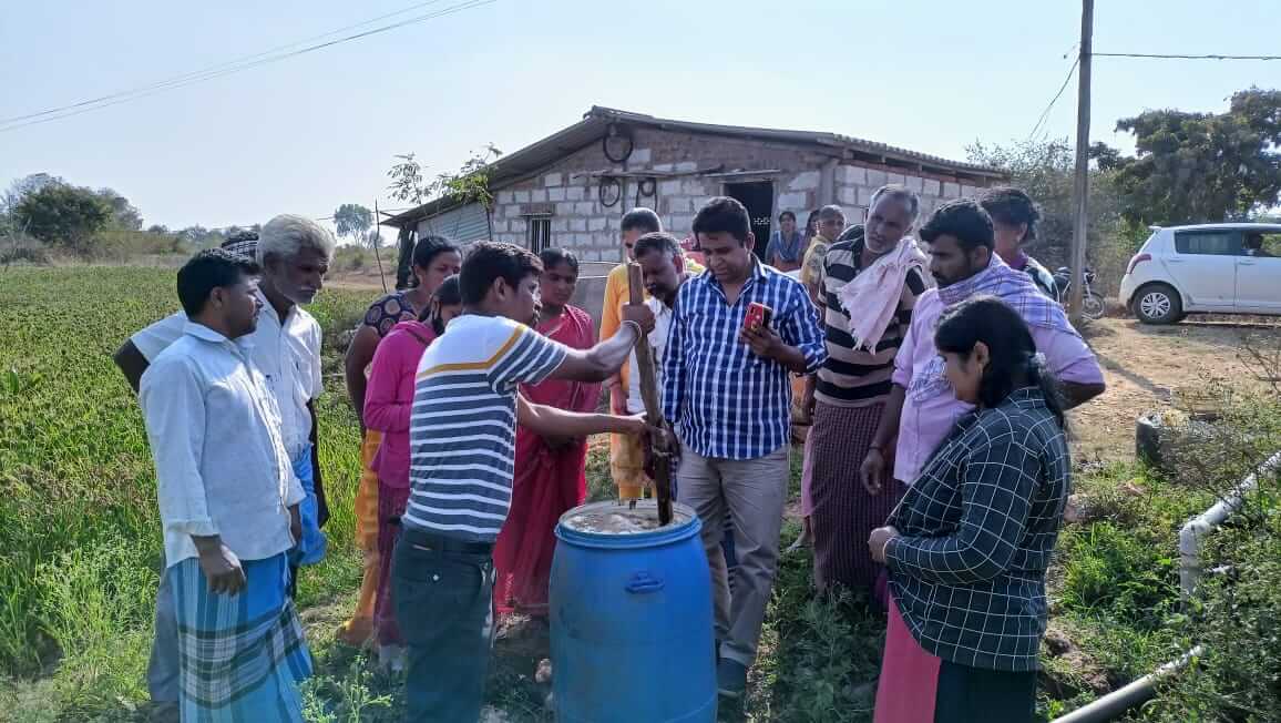 Farmers gather around a blue barrel during training on agroecological methods involving liquid manure near a rural building.