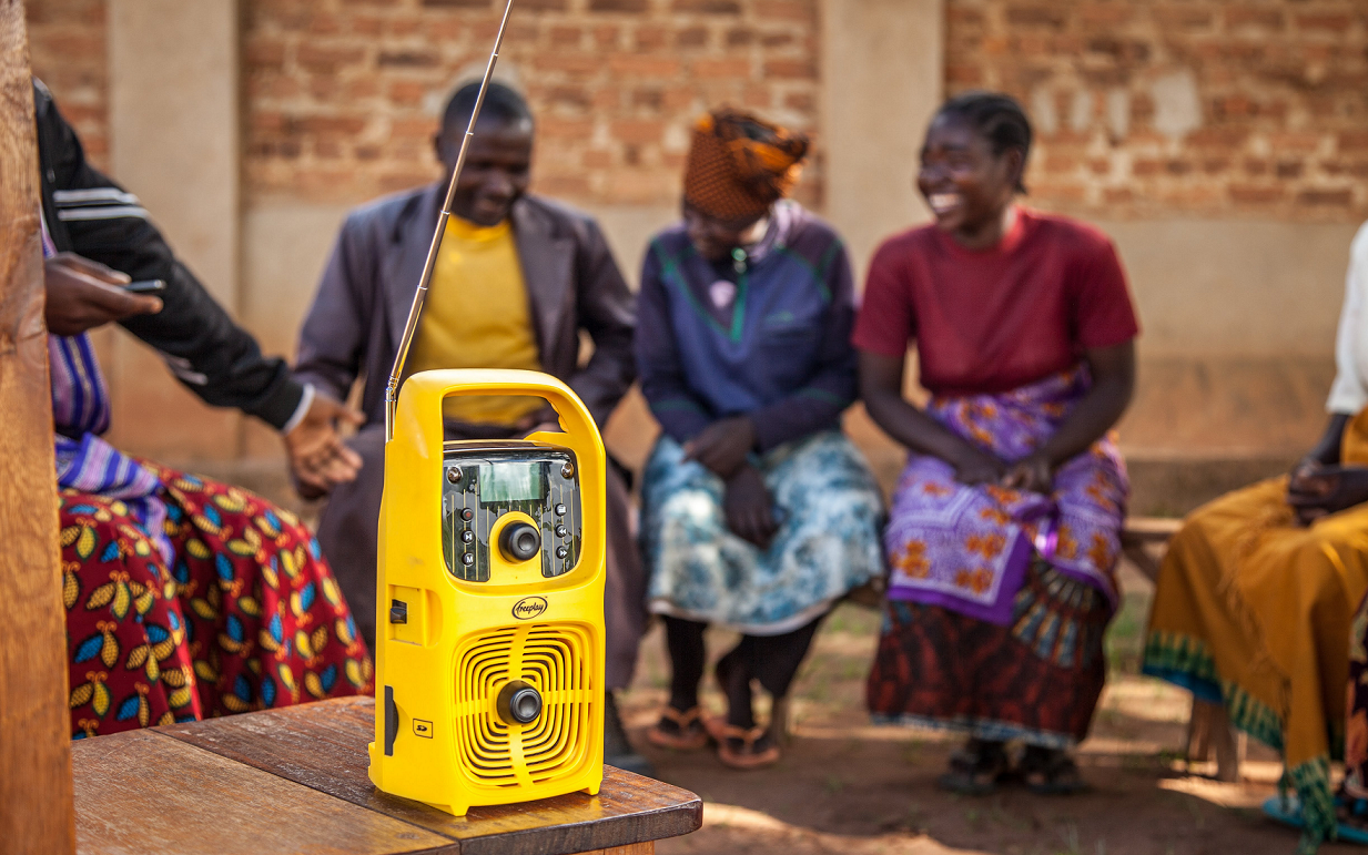 Eine Gruppe von Menschen sitzt lachend um ein gelbes Radio und hört gemeinsam zu.
