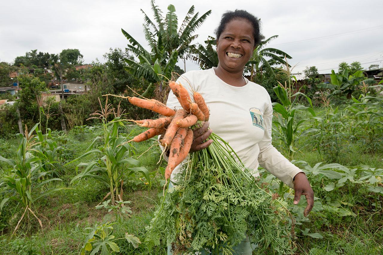 A smiling woman proudly holds freshly harvested carrots in a lush garden, showcasing her organic bounty.