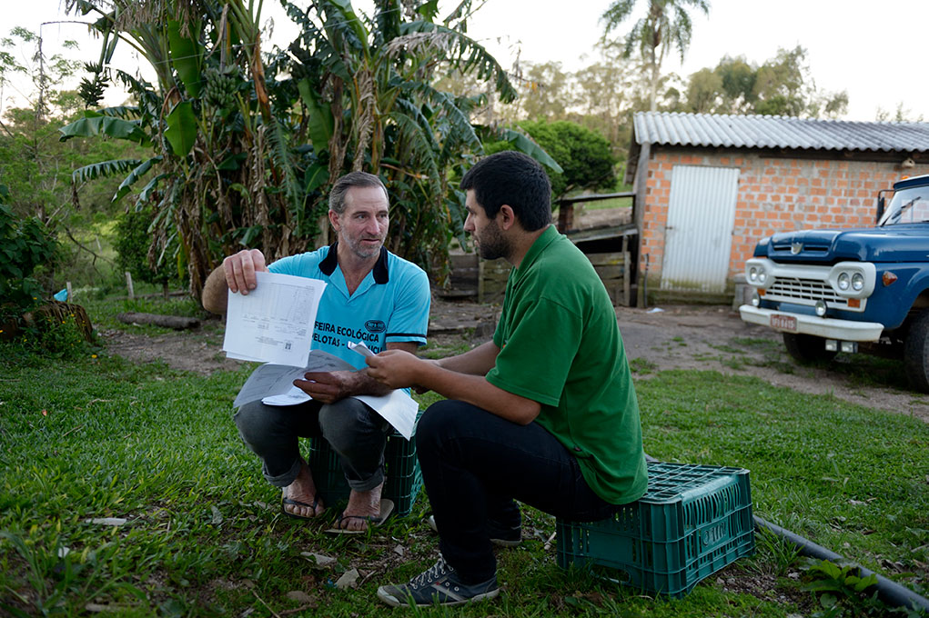 Two men discuss documents outdoors near a farm, with a vintage truck and lush greenery in the background.