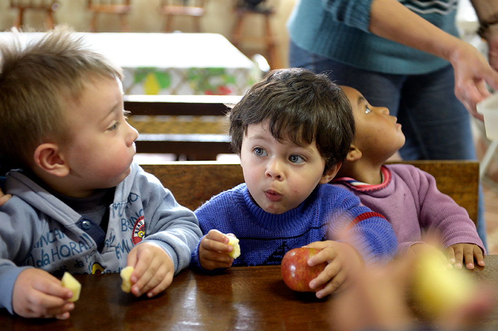 Three young children sit at a wooden table, holding apples and fruit pieces, with curious expressions.