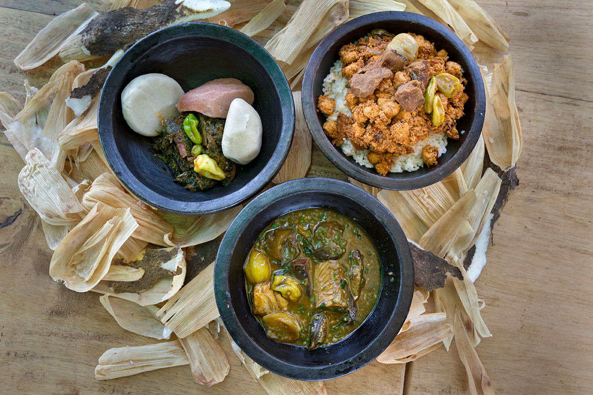 Drei Schalen mit traditionellen afrikanischen Gerichten auf Holztisch, umgeben von getrockneten Maisblättern.