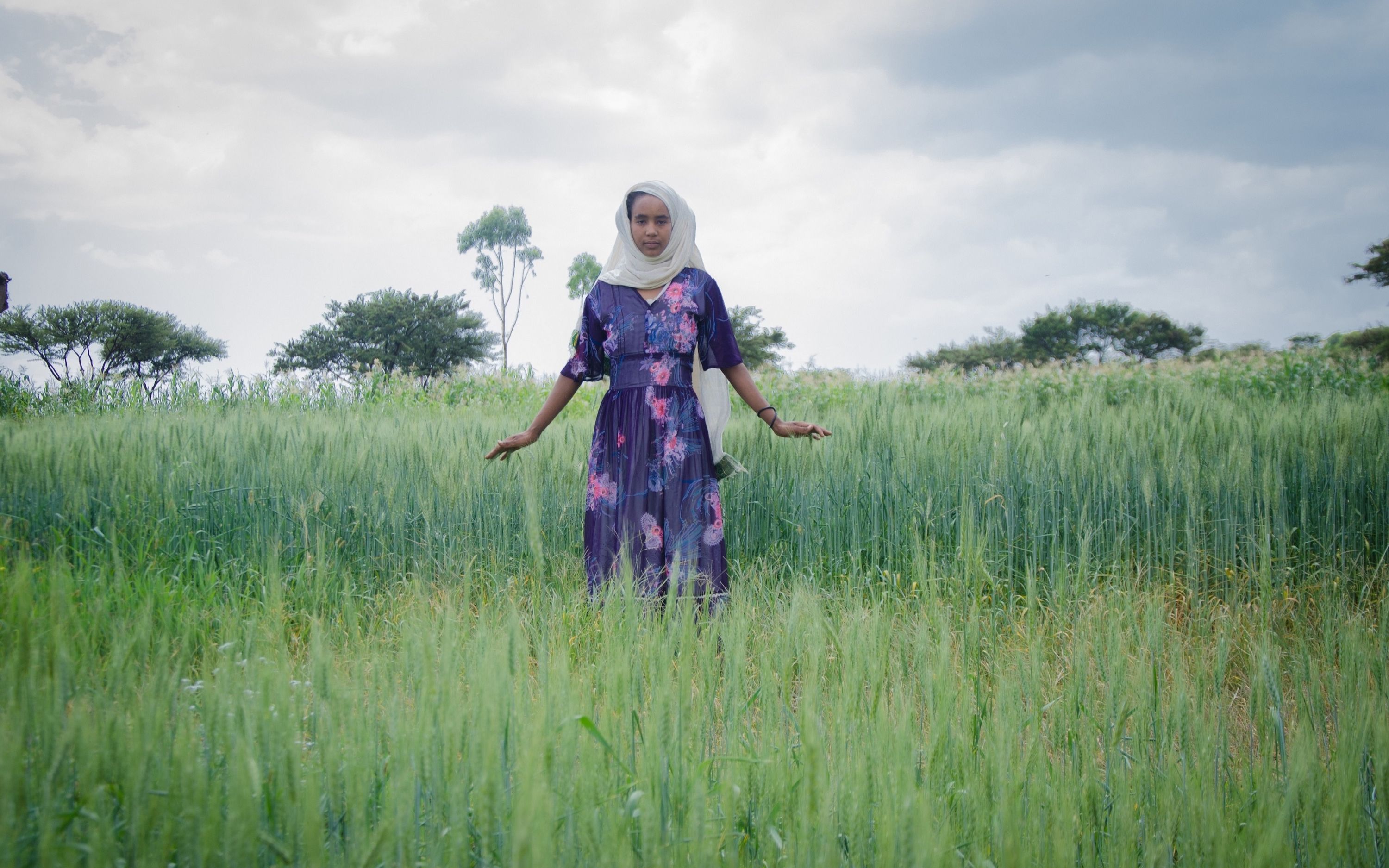 A woman in a floral dress stands gracefully in a lush green field under a cloudy sky, embodying tranquility.