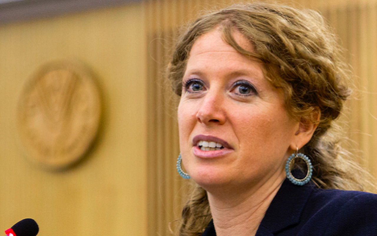 Eine Frau mit lockigem Haar und blauen Ohrringen spricht in einem formellen Raum vor einem Mikrofon.