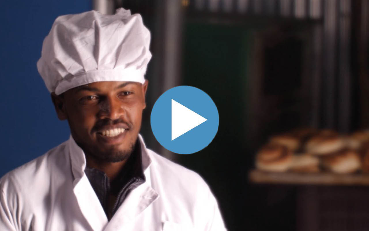A smiling baker in a white uniform stands in a bakery with freshly baked bread in the background.